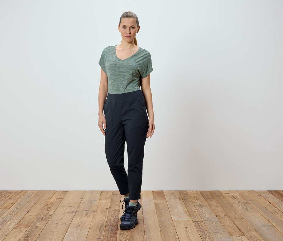 Une femme se tient devant un mur blanc sur un plancher en bois. Elle porte un t-shirt vert et un pantalon noir.
