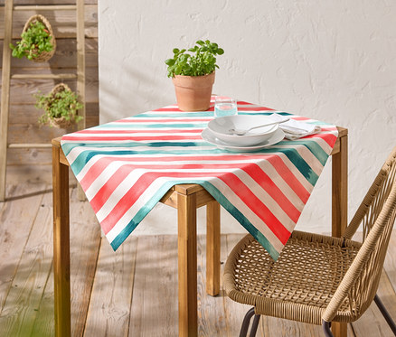 Une table avec une nappe rayée rouge, bleu et blanche, des assiettes, des couverts et une boisson est installée à l'extérieur.