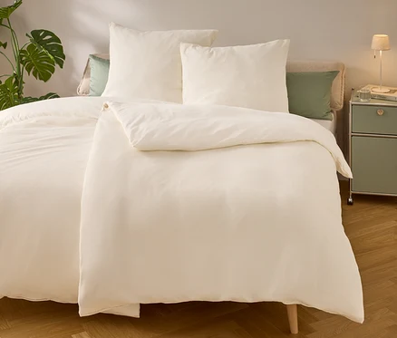 Lit avec des draps et des oreillers beige clair. Une lampe et un livre sont posés sur la table de chevet. Une plante verte est à gauche du lit.
