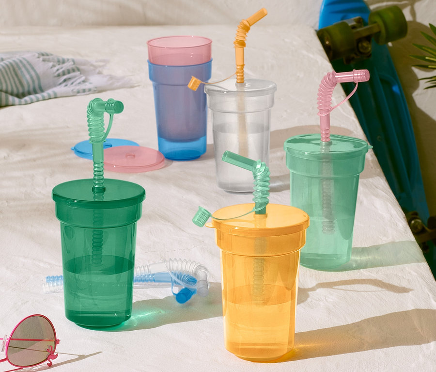 Un lot de gobelets pour enfant de couleur verte, jaune, transparente et bleue avec des pailles sur une surface blanche à côté d'un skateboard et de lunettes de soleil.