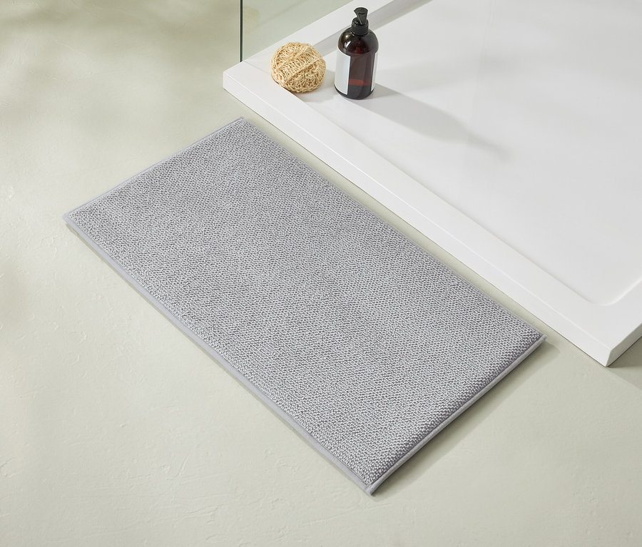 Un tapis de bain gris clair est posé à côté d'un receveur de douche blanc sur lequel reposent une bouteille marron et un luffa.