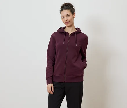 Une femme portant un chignon porte un sweat à capuche bordeaux zippé et un pantalon noir sur fond blanc.