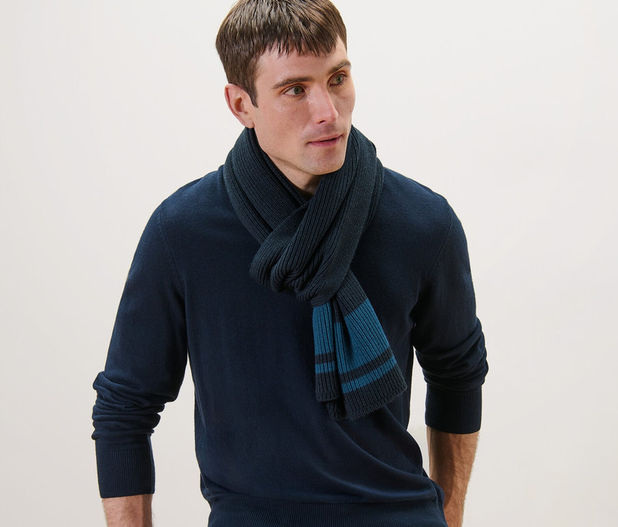 Un homme portant une écharpe en maille, un pull bleu marine et un jean, appuyé sur une chaise.