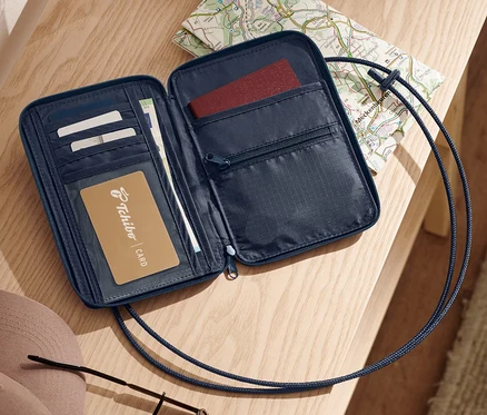 Une pochette de poitrine de sécurité bleue ouverte repose sur une surface en bois clair à côté d'une carte, de clés, d'un chapeau et de lunettes de soleil.