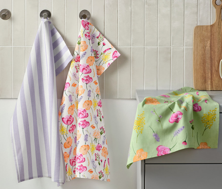 Trois torchons sont accrochés au mur : un torchon rayé violet et blanc, un avec un motif floral et un sur le comptoir. À côté se trouvent une planche à découper et une tasse avec des ustensiles de cuisine. 3 linges de cuisine