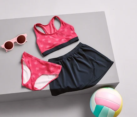 Des lunettes de soleil roses, un slip de bikini à motif de palmiers, une brassière de sport et une jupe bleu marine sont posés sur une surface grise, avec un ballon de volley multicolore.
